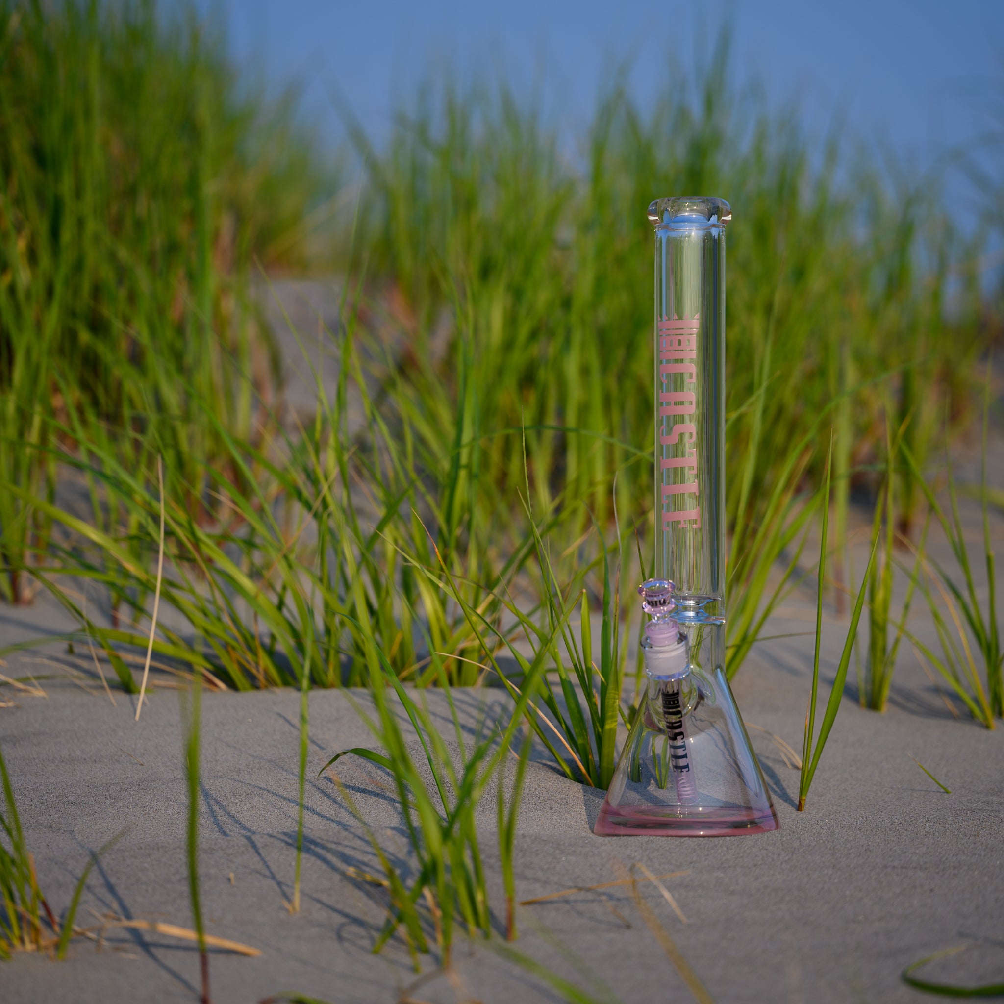 A clear glass beaker bong with pink accents and 'CASTLE' branding on the neck, standing upright on a sandy beach among green grass. The sunlight highlights the smooth glass, with a serene blue sky in the background.