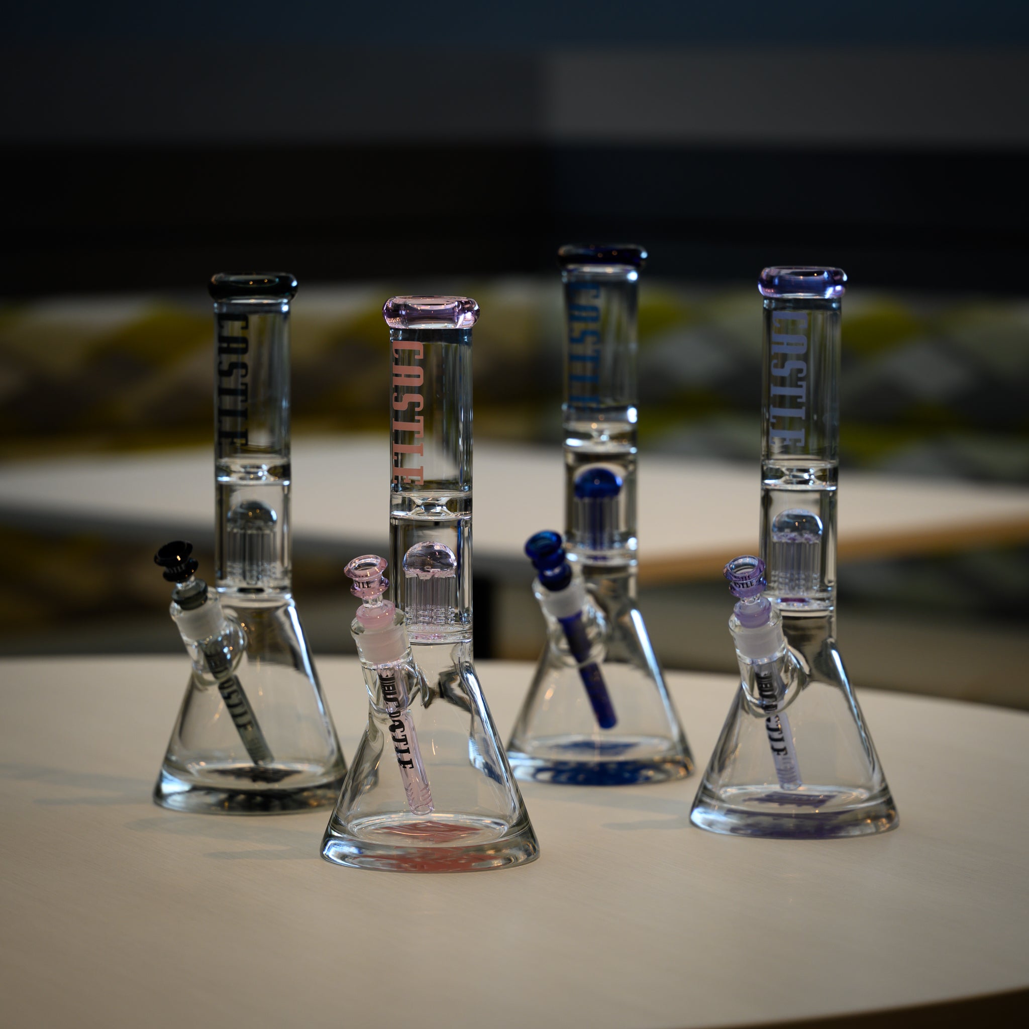 A set of four clear glass beaker bongs with colored accents in black, pink, blue, and purple, displayed on a white table. Each bong features the 'CASTLE' branding on the neck and matching downstems and bowls, arranged in a stylish indoor setting.