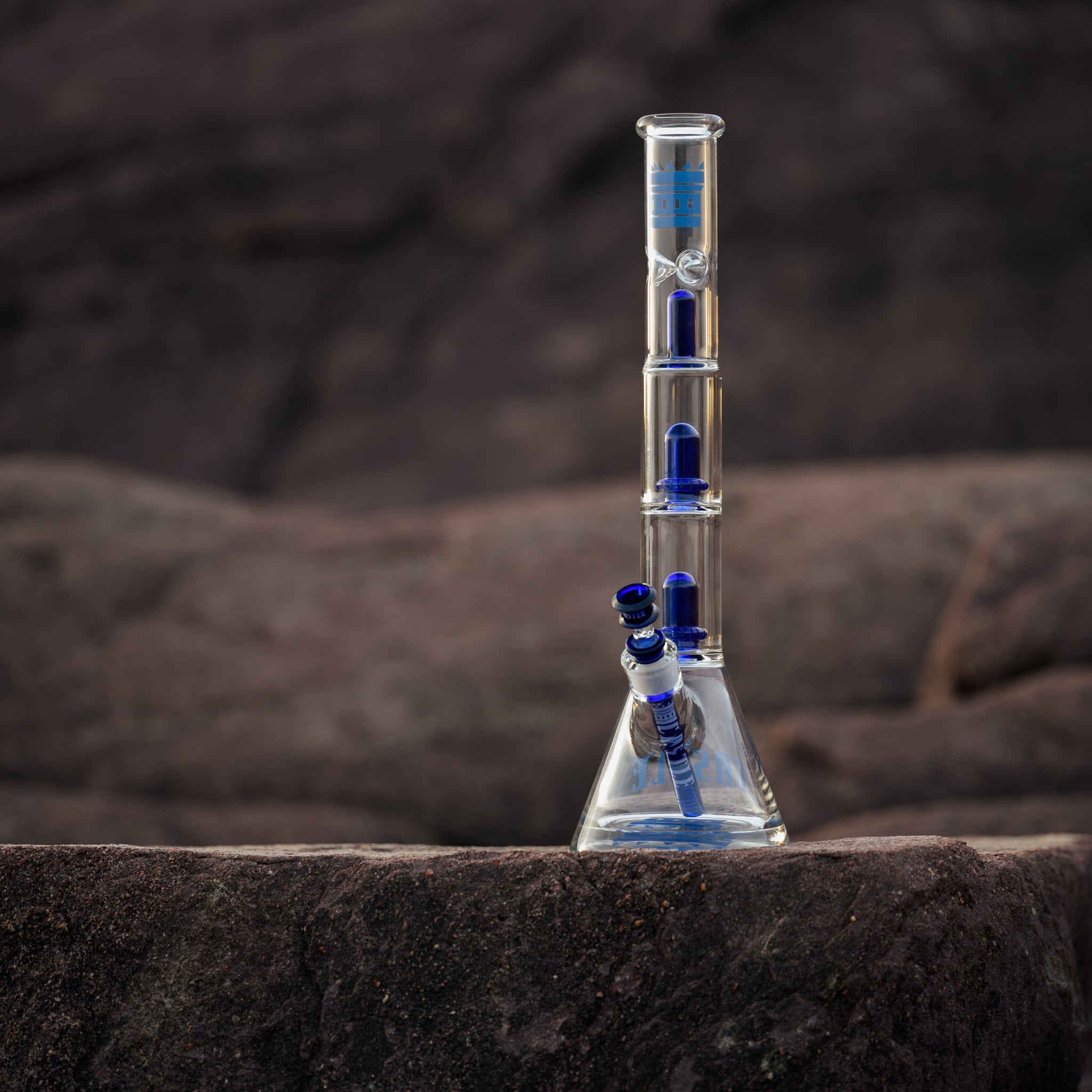 18" intricate glass bong with bold blue accents stands on a rugged rock surface, set against a backdrop of earthy stone formations. The piece features multiple percolation chambers, each equipped with matching blue percs for enhanced filtration. The downstem and bowl also share the deep blue color, complementing the overall design. The natural, muted tones of the rocky landscape contrast with the bong’s sleek transparency and vibrant accents, highlighting its craftsmanship and high-performance functional.