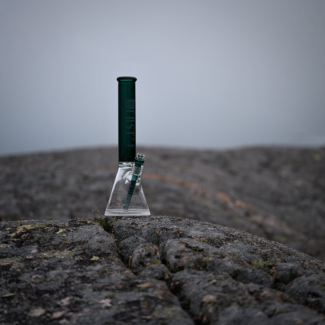 A glass beaker bong with teal colored glass, including the neck and downstem, featuring the "Castle" logo vertically etched on the neck. The bong is placed on a rugged, rocky surface with a foggy, misty background, creating a moody and serene atmosphere. The overcast sky and muted tones of the scene enhance the contrast between the sleek, modern design of the bong and the natural, textured rock.