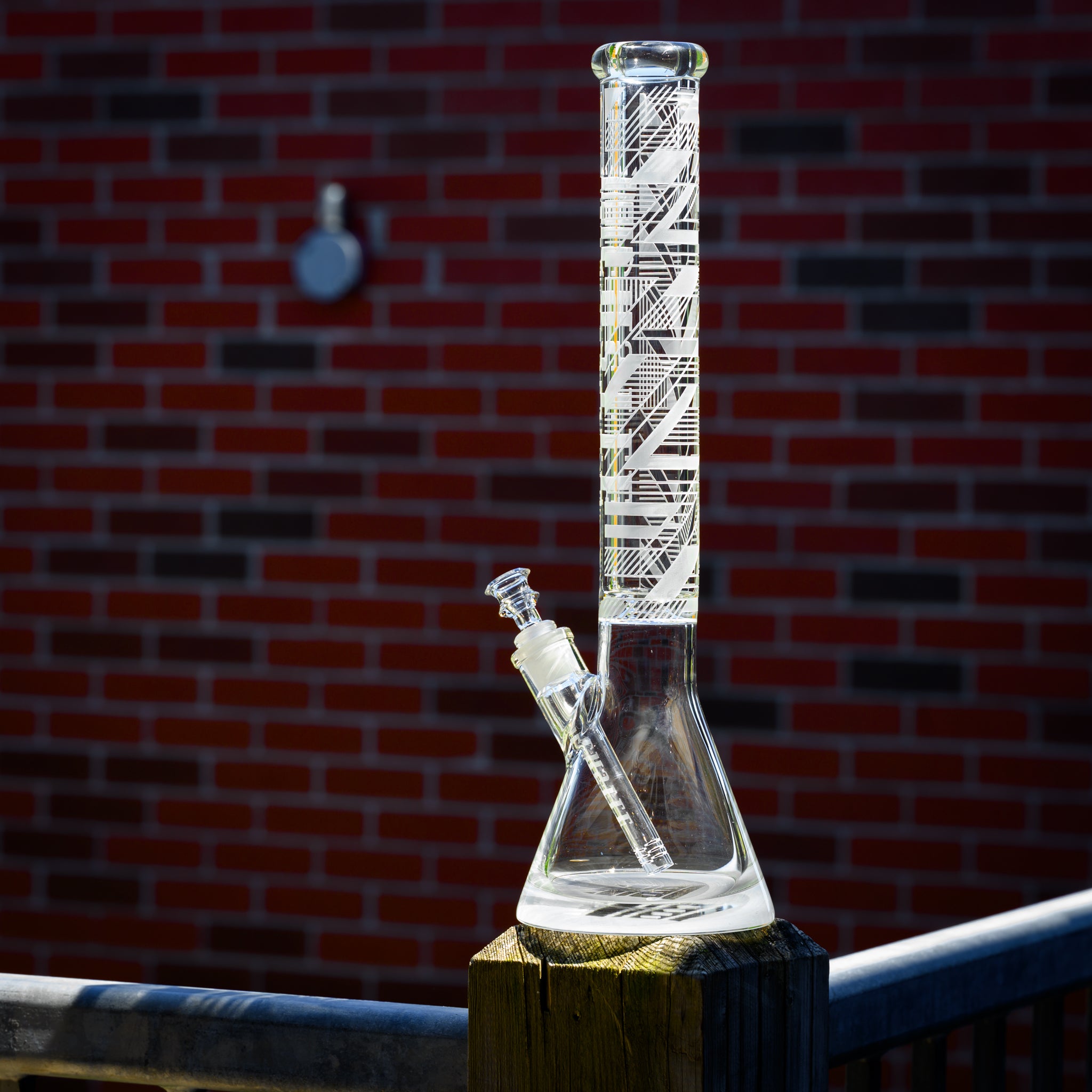 Clear glass beaker bong with intricate geometric etching on the neck, placed on a wooden post. The background features a red brick wall with a blurred metal fixture, adding contrast to the sharp details of the glass.