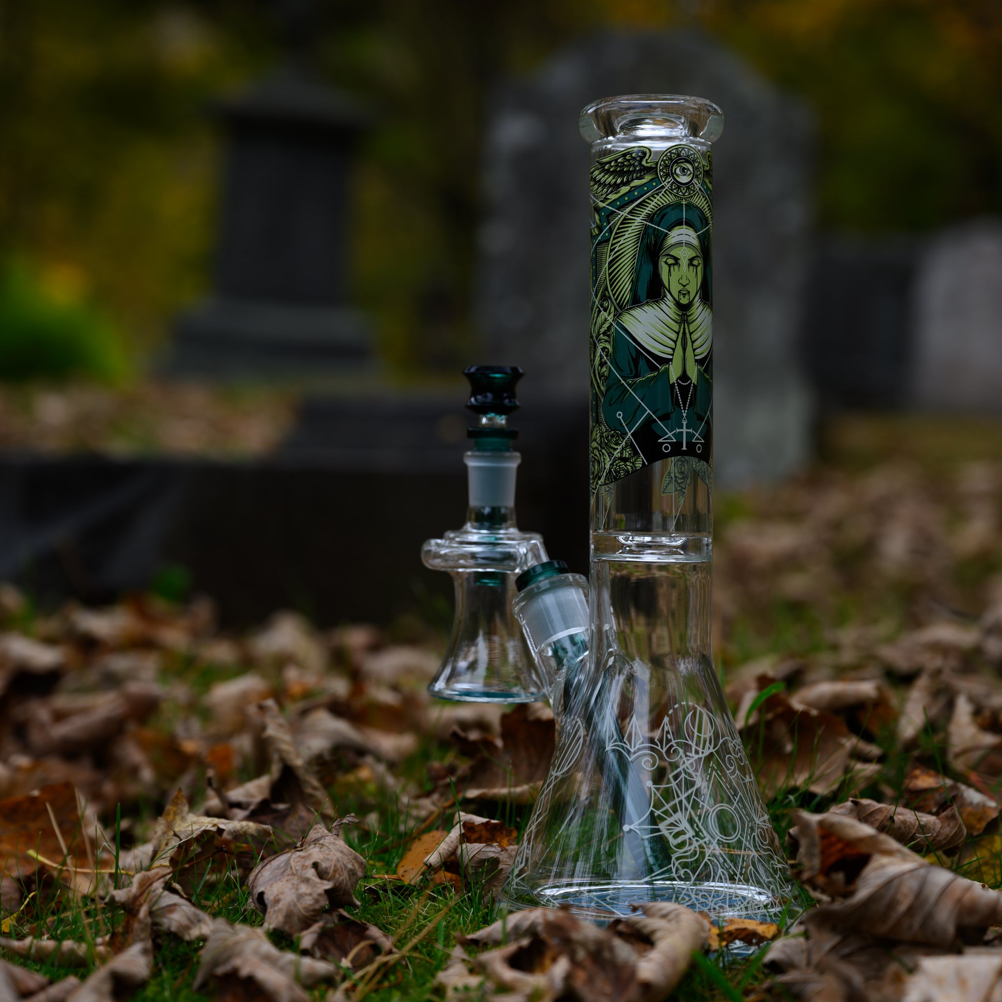 A gothic-inspired glass bong stands among fallen autumn leaves in an eerie graveyard setting, surrounded by weathered tombstones. The neck features a haunting, medieval-style illustration of a regal, mysterious woman framed by intricate geometric patterns. The beaker base is adorned with celestial etchings, adding to the mystical aesthetic. A matching glass recycler attachment enhances its functionality and design. 