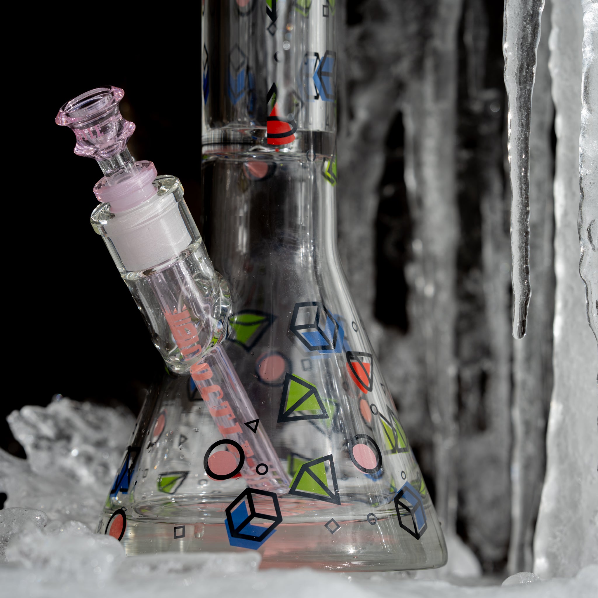 A close-up of a clear glass beaker bong with colorful geometric shapes, featuring a pink-accented bowl and downstem. The bong is set against a backdrop of ice and icicles, creating a contrast between the vibrant design and the frozen environment.