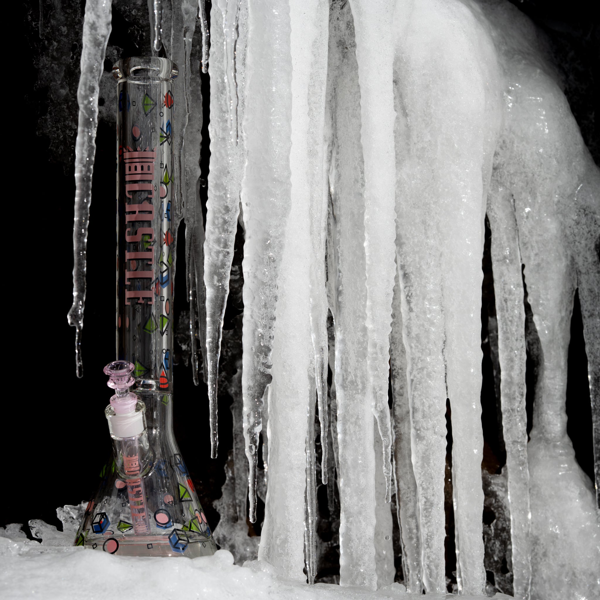 A clear glass beaker bong with colorful geometric designs and pink accents, standing against a backdrop of large icicles. The frozen setting highlights the intricate details of the glass, creating a striking contrast between the bong and the ice formations.
