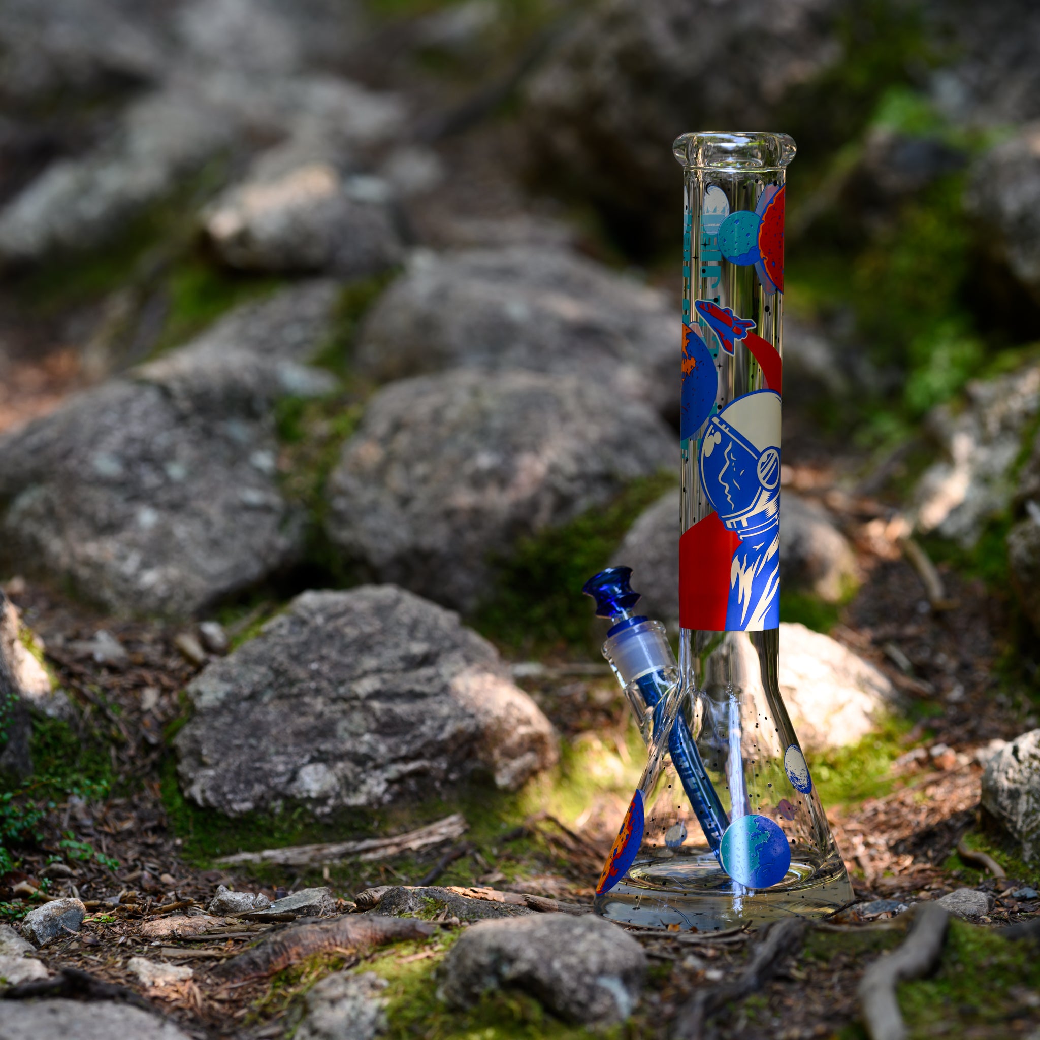 A glass bong with a vibrant space-themed design stands among moss-covered rocks in a sunlit forest. The neck features bold blue, red, and white illustrations of planets, astronauts, and cosmic elements, creating a striking contrast against the clear glass. The cobalt blue bowl and downstem complement the design, adding depth and cohesion to the aesthetic. Dappled sunlight filtering through the trees enhances the natural setting, emphasizing the craftsmanship and adventurous spirit of the piece.