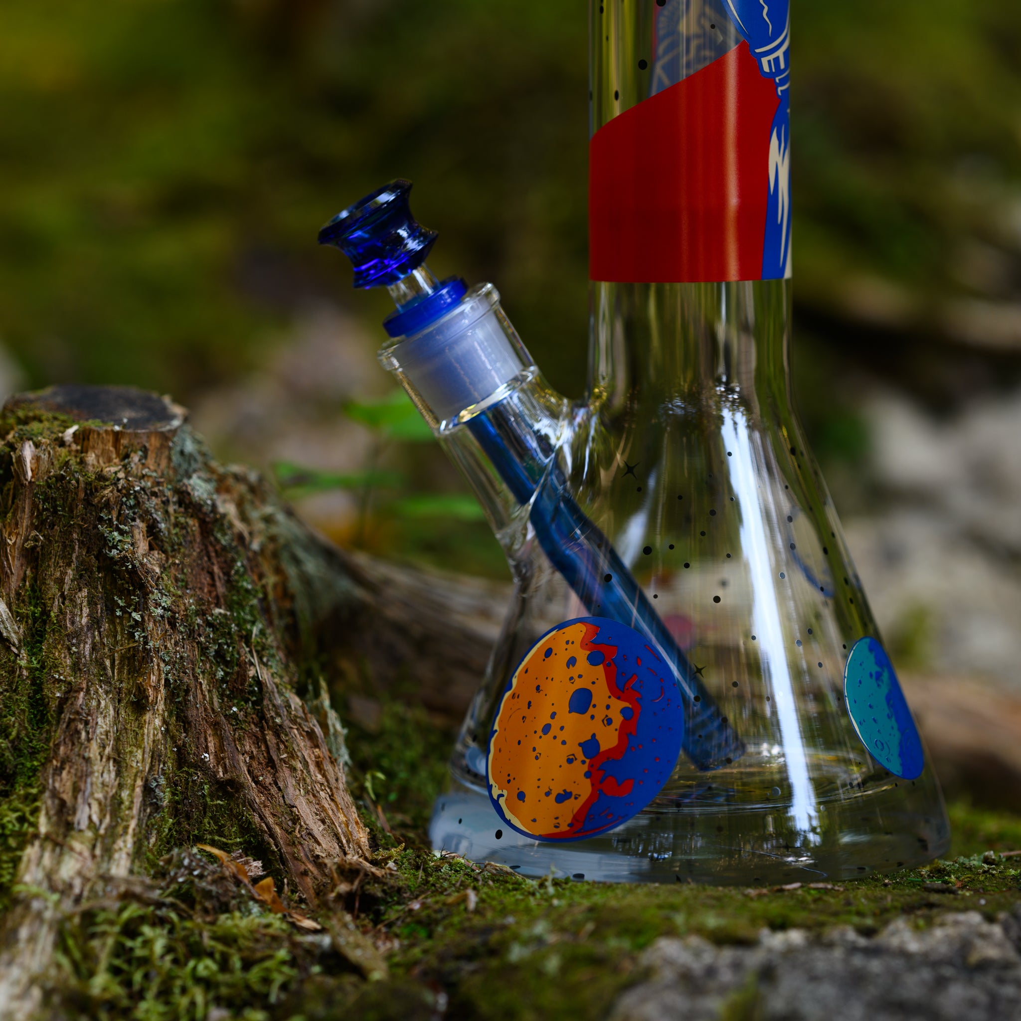 A close-up of a space-themed glass bong resting on a mossy forest floor near a weathered tree stump. The beaker-style base features colorful planetary illustrations, with a bright orange and blue cratered planet prominently displayed. The rich cobalt blue downstem and matching bowl complement the cosmic design, adding depth to the piece. The natural surroundings, with soft green moss and earthy textures, contrast beautifully with the sleek transparency of the glass, emphasizing craftmanship.