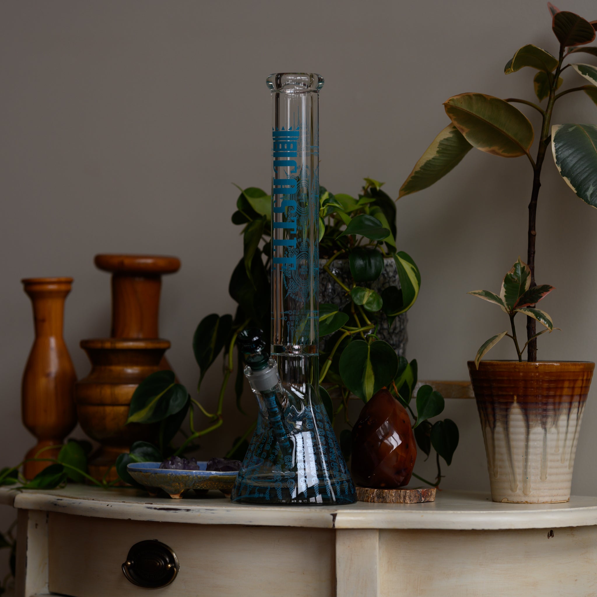 Tall clear glass water pipe with blue decorative text and artwork, displayed on a wooden table near green houseplants and small decorative items.